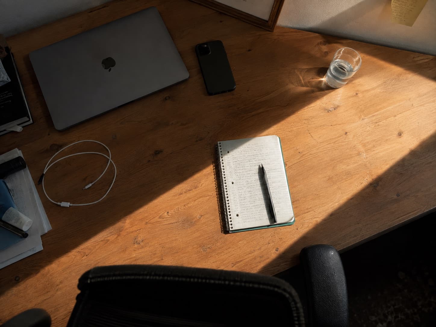 Mesa com o notebook fechado, o celular virado para baixo e um caderno aberto, iluminados por um facho de sol